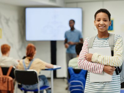 Waist up portrait of smiling teenage girl holding books and looking at camera in school classroom, copy space